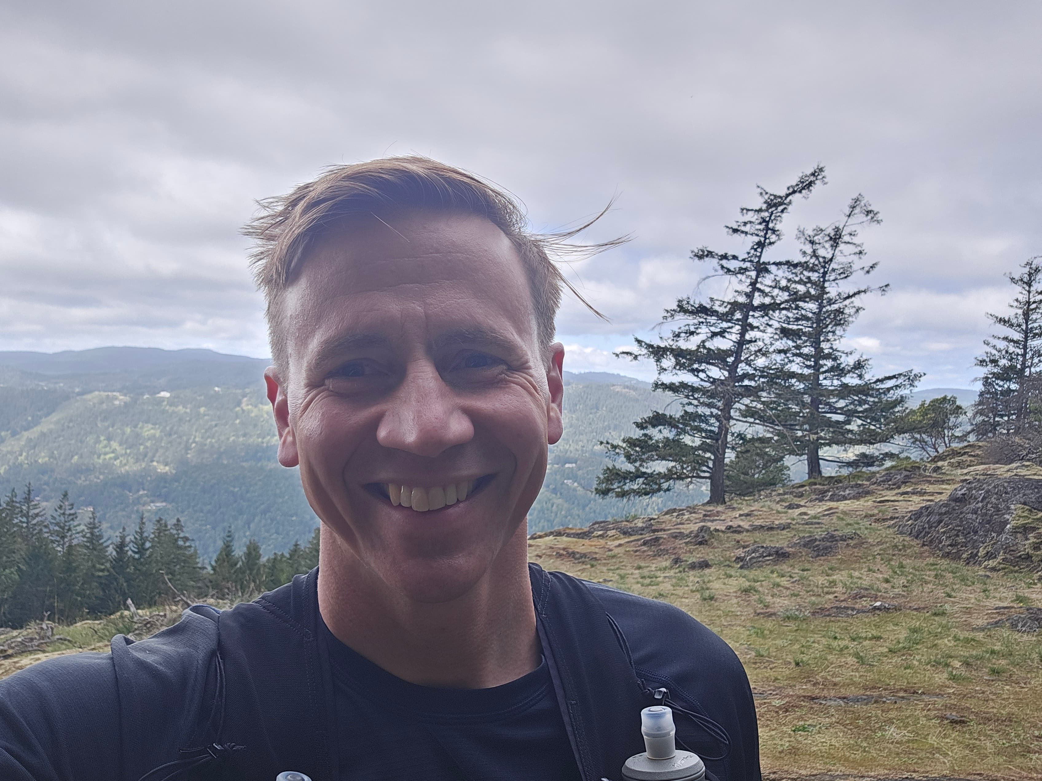 Portrait of JME Maxwell — close-up smile, mountains and forest in the background, wind in his hair.
