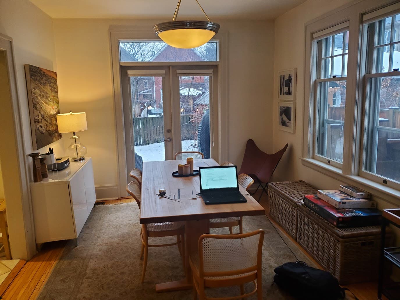 A laptop open on a wooden dining table in a home, French doors onto a snow-covered backyard, warm pendant light overhead, board games stacked on a bench by the window. The office that wasn't an office.