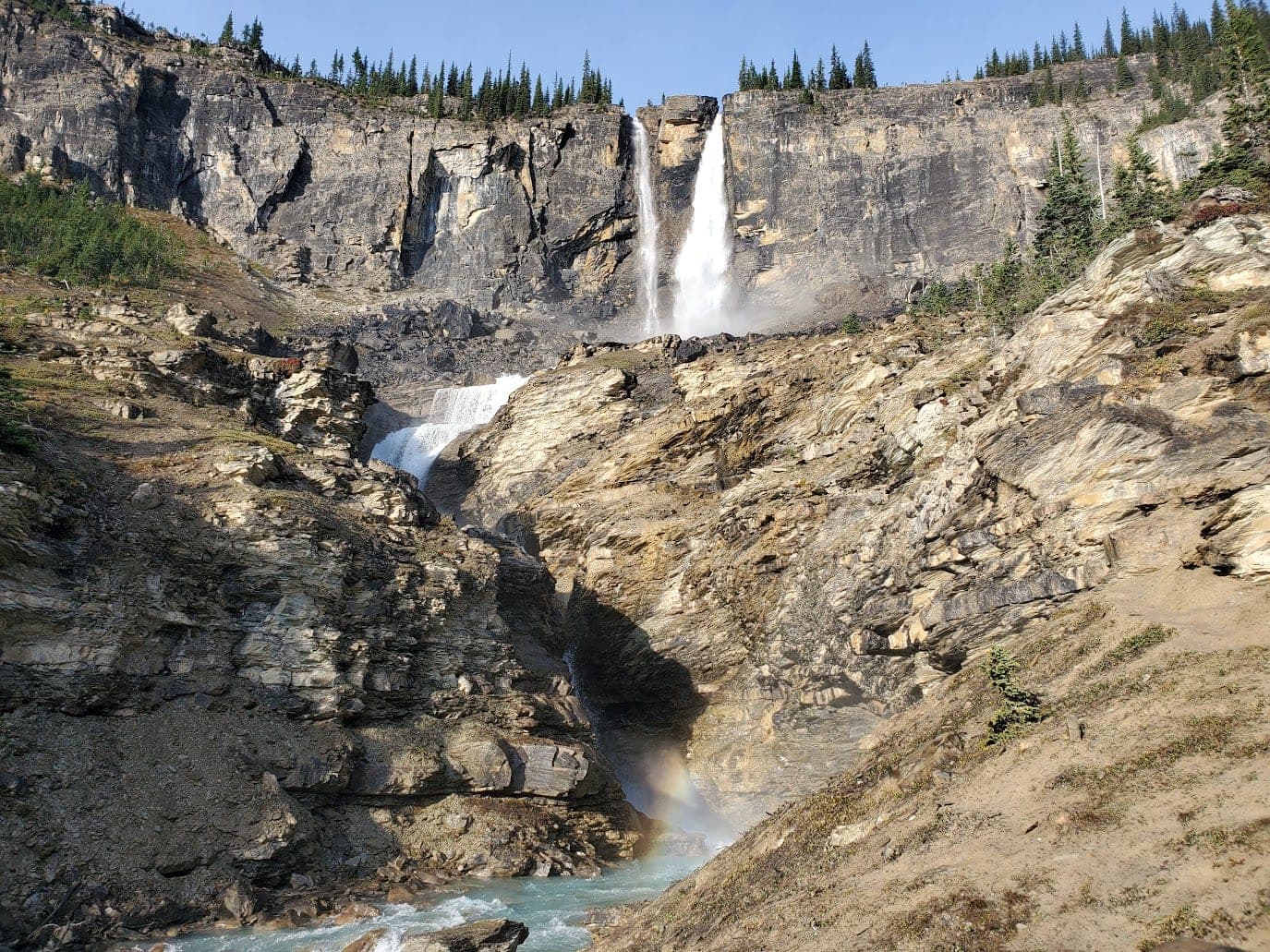 A tall twin waterfall pouring off a cliff band in the Rockies, turquoise river below.